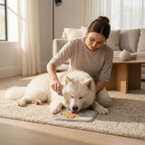 Nuzzo dog lick mat with textured surface being used by a white Samoyed dog while a woman grooms it in a bright room.